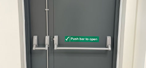A grey double fire escape door with horizontal push bars and a green sign that reads 