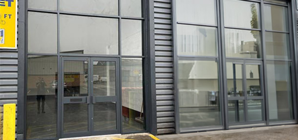 A commercial building exterior featuring a modern aluminum-framed glass shopfront entrance on the left and an industrial loading bay with a roller shutter door on the right. The facade is clad in grey metal panels, with yellow safety bollards and protective barriers positioned near both the entrance and the loading area.
