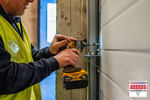 A closeup image of a person in a high-Viz jacket drilling into a sectional door with a portable, hand-held drill.