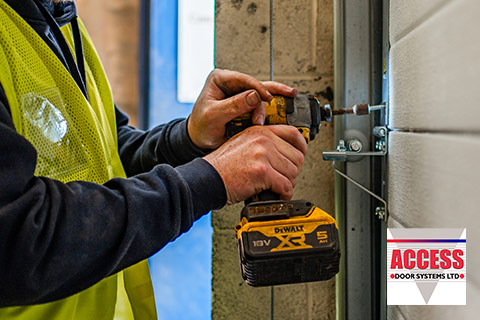 A closeup image of a person in a high-Viz jacket drilling into a sectional door with a portable, hand-held drill.