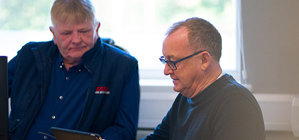 2 men sitting at a desk and looking down at an electronic tablet / ipad